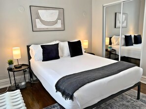 A modern hotel room with a large bed, two bedside tables, a mirror, and a wall-mounted abstract art piece. - Smart & Stylish 2-Bedroom Unit (Chicago)