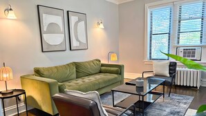 Una sala de estar moderna con un sofá verde, un sillón de cuero, una mesa de centro de vidrio y una lámpara de pie. Hay dos luminarias fijadas a la pared y un radiador cerca de la ventana.