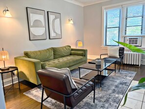 A modern living room with a green sofa, a leather chair, a glass coffee table, and a floor lamp. There are two wall-mounted light fixtures and a radiator near the window. - Smart & Stylish 2-Bedroom Unit (Chicago)