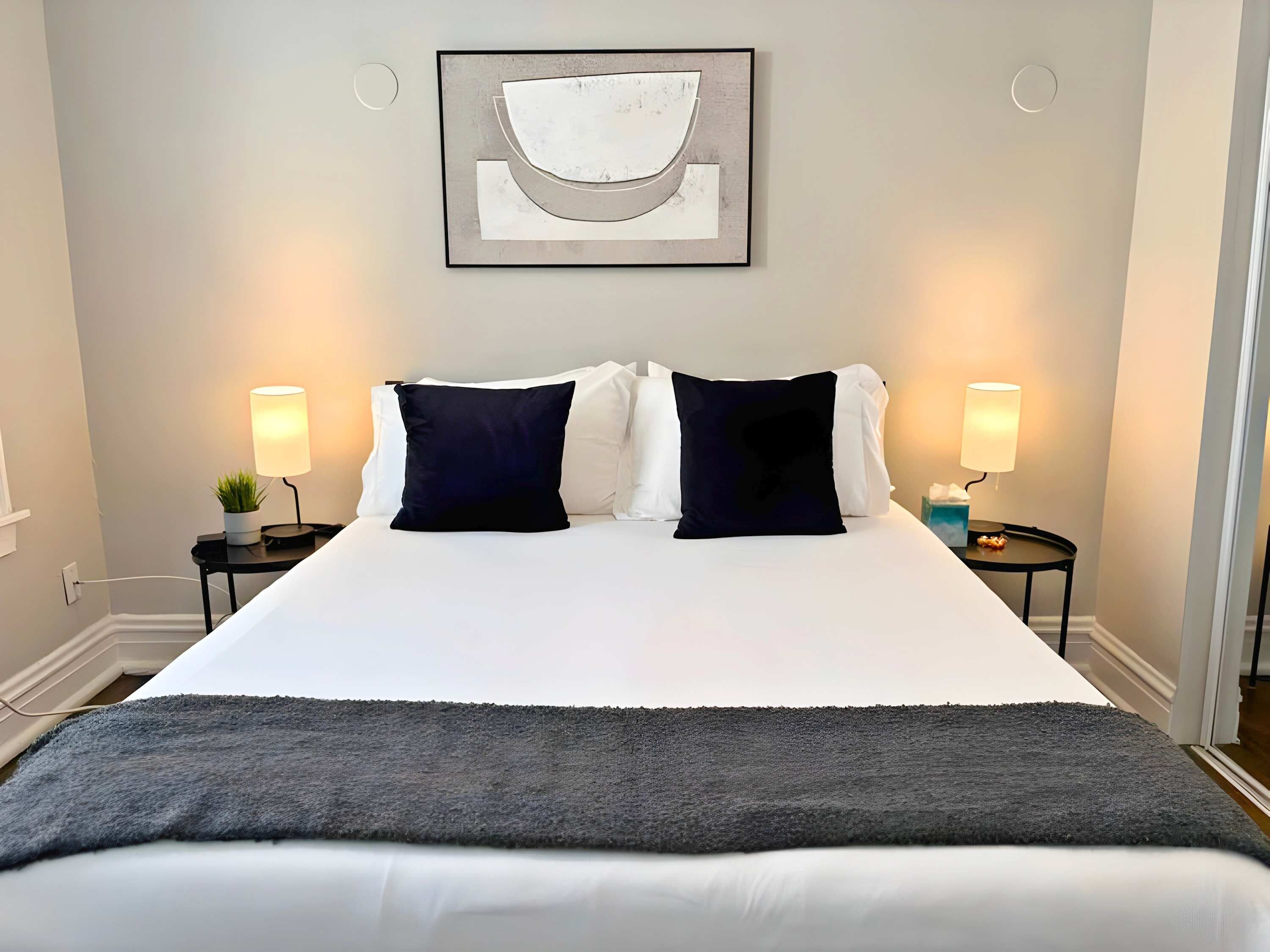 A neatly made bed with white linens and two dark pillows, flanked by two bedside tables with lamps, a framed abstract artwork above the bed, and a grey throw blanket at the foot of the bed.