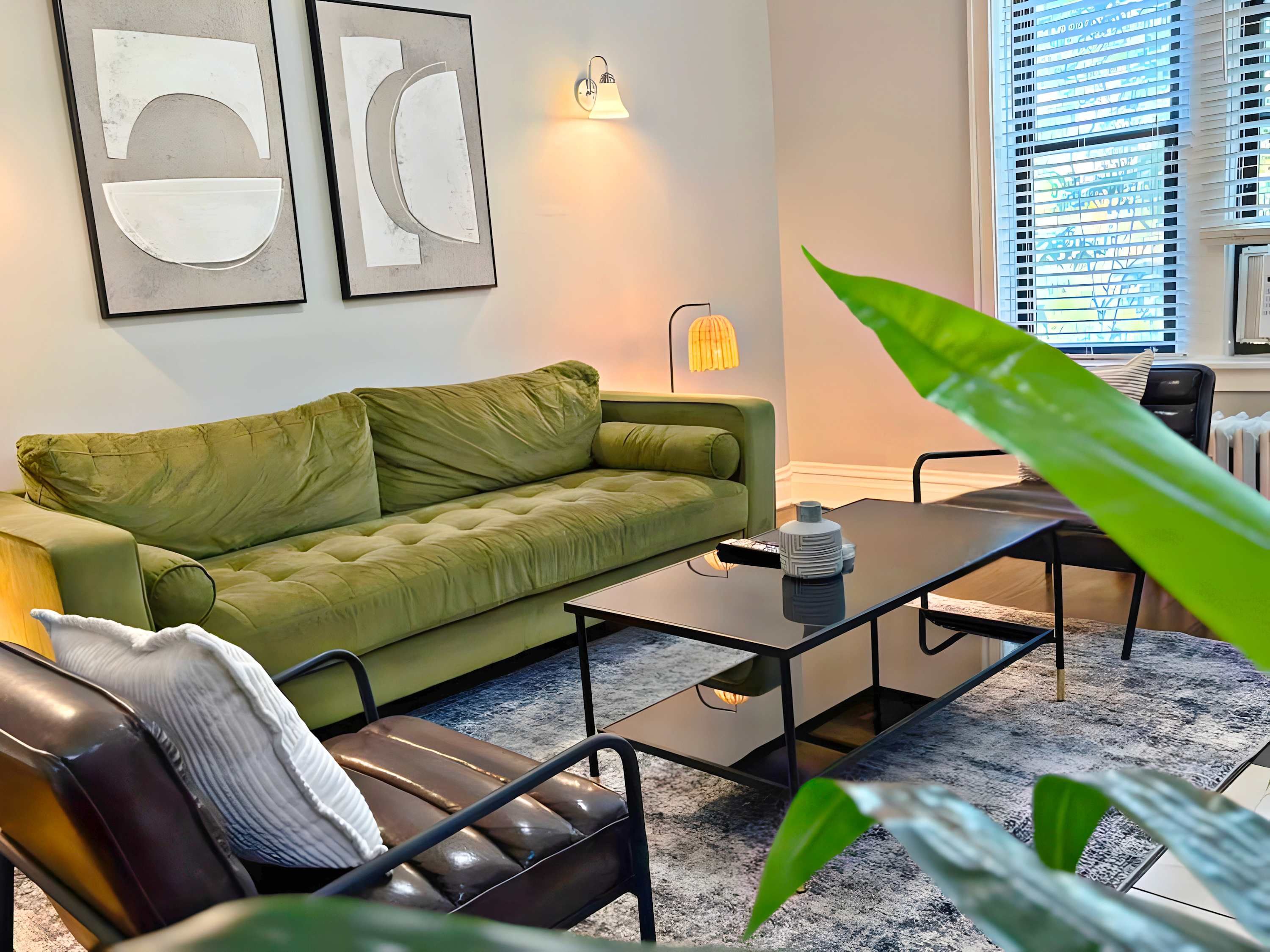 A modern living room with a green sofa, a black leather chair, and a coffee table. There are abstract wall art pieces and a wall-mounted lamp.
