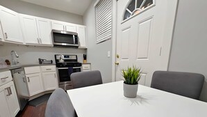Cocina moderna con armarios blancos, microondas, estufa y horno. Hay una mesa de comedor con dos sillas y una planta en maceta.
