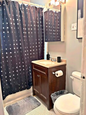 A bathroom with a dark curtain featuring a white crescent moon pattern, a wooden vanity with a beige countertop, a white toilet, and a small mirror above the sink. - Smart & Stylish 2-Bedroom Unit (Chicago)