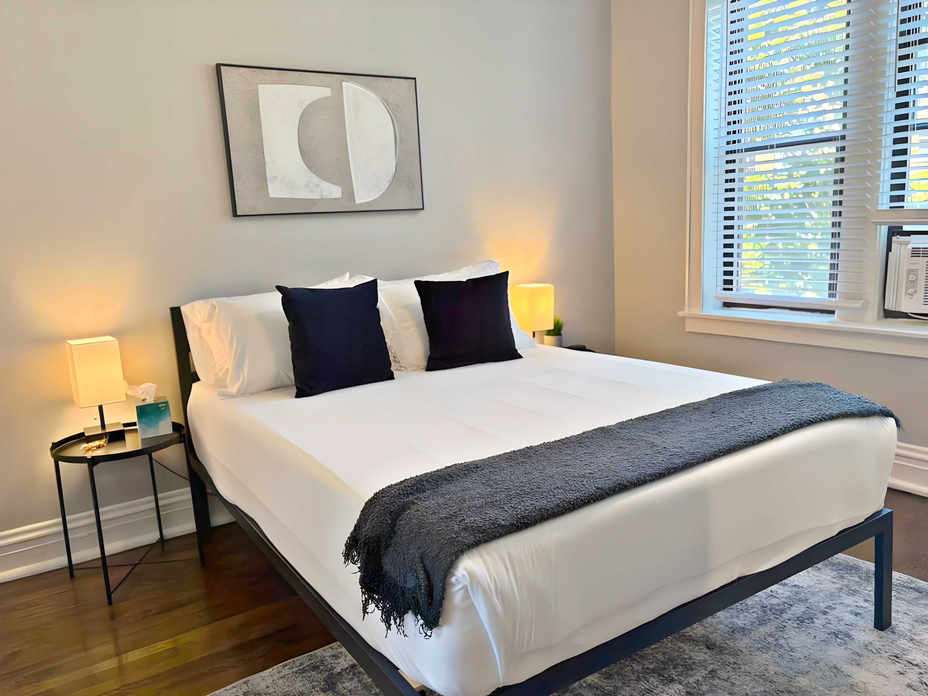 Een opgemaakt bed met een donker hoofdbord, twee nachtkastjes met lampen, een lijst met abstracte kunst aan de muur en een raam met jaloezieën.