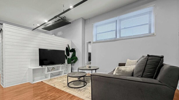 En moderne stue med et fladskærms-tv, en grå sofa, et rundt bord og en potteplante.