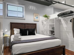 Een keurig opgemaakt bed met witte en zwarte kussens, een bedlampje en een klein tafeltje met een lamp.