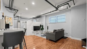 En moderne stue med en grå sofa, et spiseområde med et hvidt bord og stole, samt et køkkenområde med et fjernsyn og en aircondition.