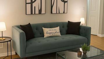 A teal sofa with a cushion that reads "THIS IS OUR happy place," flanked by two black pillows. Above the sofa, there are two abstract wall art pieces. A glass coffee table with a plant is in front of the sofa.