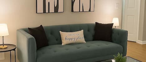 A teal sofa with a cushion that reads "THIS IS OUR happy place," flanked by two black pillows. Above the sofa, there are two abstract wall art pieces. A glass coffee table with a plant is in front of the sofa.