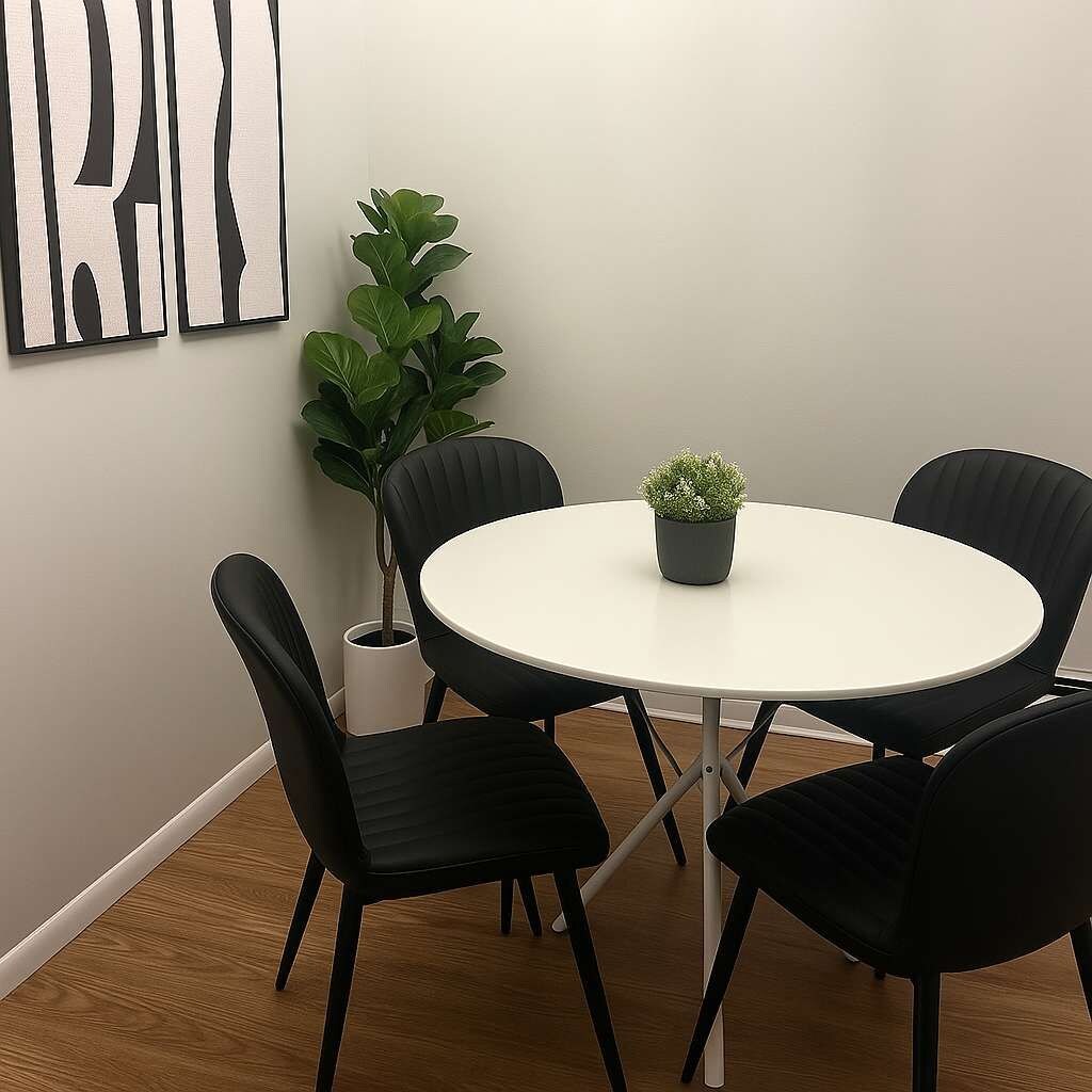 Uma mesa redonda com quatro cadeiras pretas, uma planta em vaso e uma obra de arte abstrata na parede.