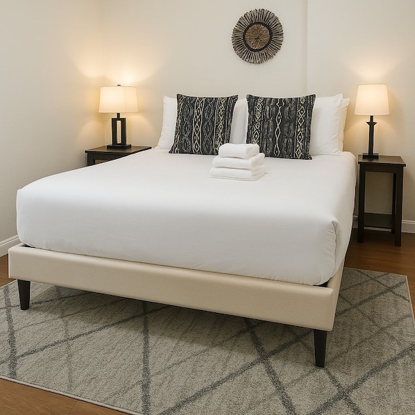 Un lit bien fait, avec une literie blanche, deux coussins à motifs et une serviette blanche pliée. On y trouve deux tables de chevet avec des lampes, un élément décoratif mural et un tapis à motifs sur le sol.