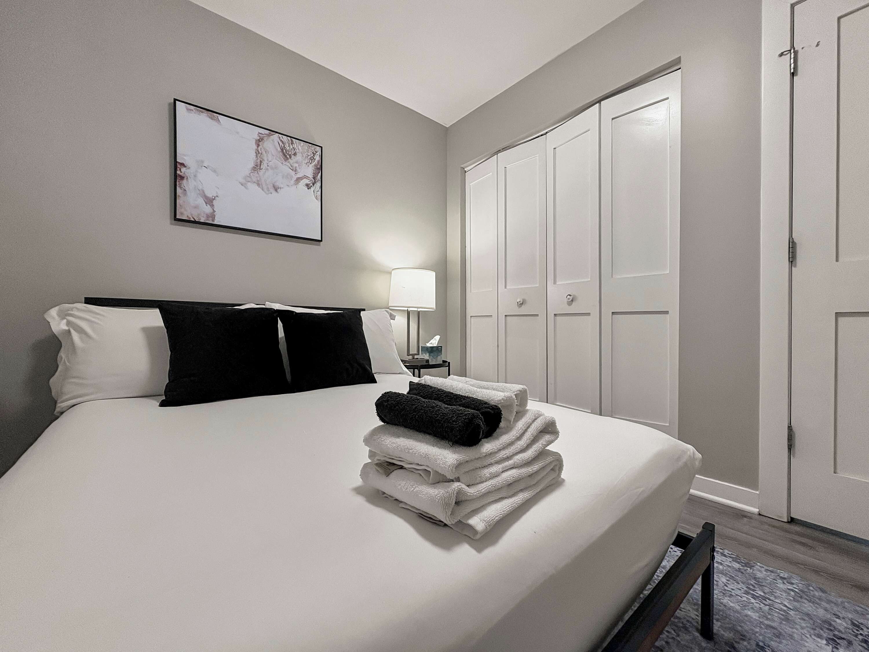 Een hotelkamer met een bed, nachtkastje, lamp, een stapel opgevouwen handdoeken en een kledingkast.