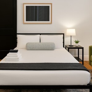 Ein ordentlich bezogenes Bett mit schwarzem Kopfteil, weißen Kissen und einer grauen Decke. Über dem Bett hängt ein gerahmtes Bild, neben dem Bett steht ein kleiner Tisch mit einer Lampe und einer Pflanze und in der Ecke ein grüner Sessel.