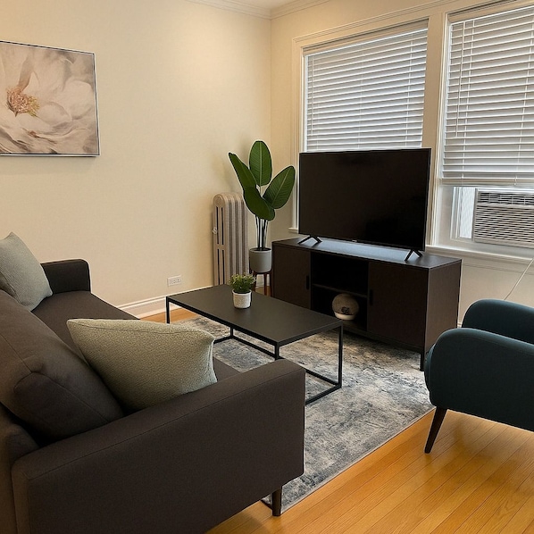 A modern living room with a sofa, a coffee table with a plant, a flat-screen TV, and a radiator. - Chic 2BR Haven Near Irving Park (Chicago)