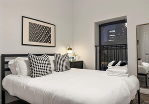 A neatly made bed with white bedding and black and white patterned pillows, a framed artwork on the wall, a bedside lamp, and a window with a view of buildings. - Modern Comfort: 1BR Unit w/ Laundry (Chicago)