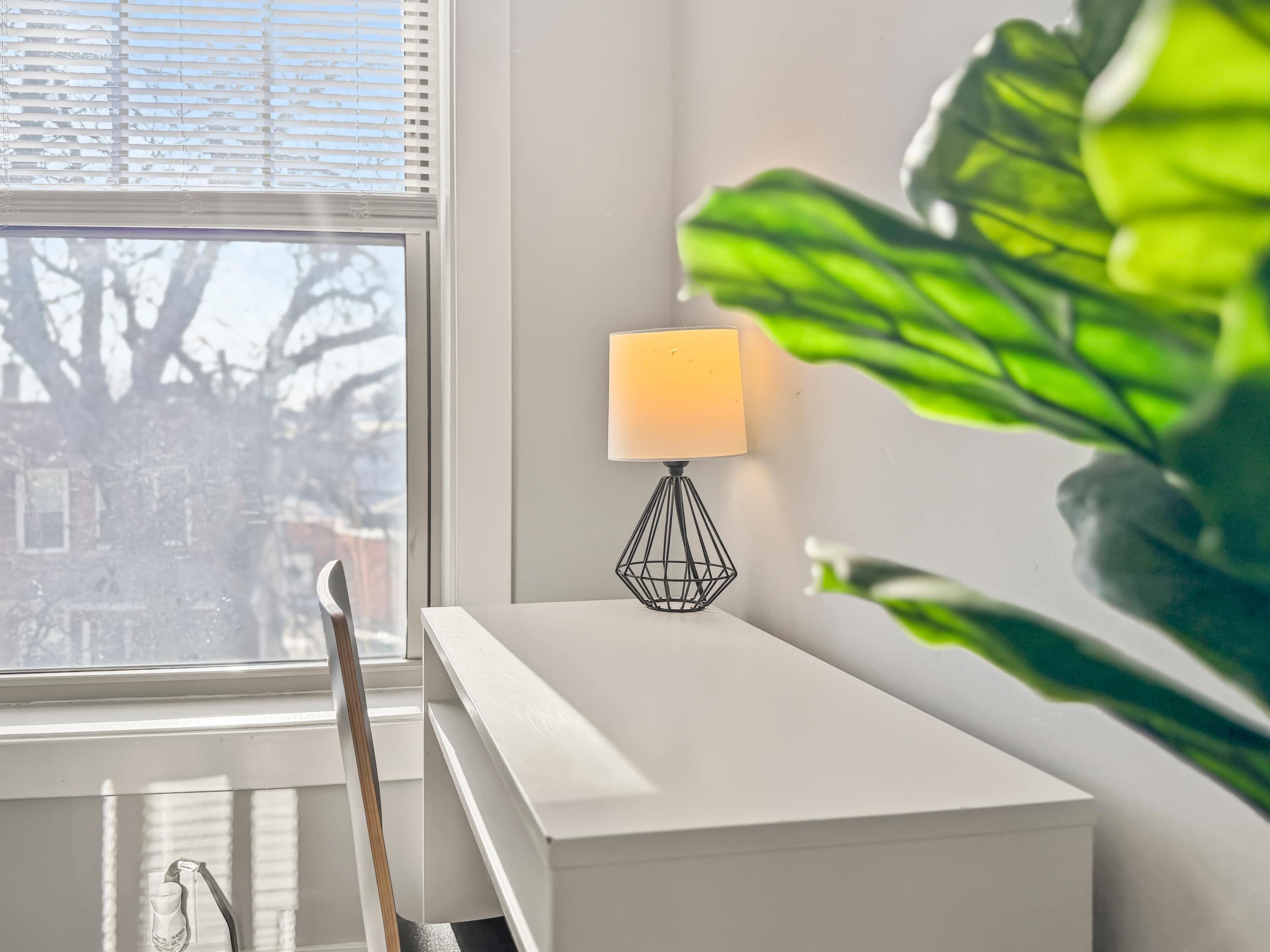 Ein weißer Nachttisch mit einer geometrischen schwarzen Lampe, ein Fenster mit Jalousien und eine große grüne Pflanze.