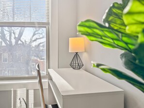 A white bedside table with a geometric black lamp, a window with blinds, and a large green plant.