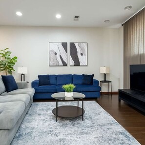 Uma sala de estar moderna com um sofá azul, um sofá cinza e uma TV. Há duas luminárias fixadas na parede e uma planta sobre uma mesa de centro.