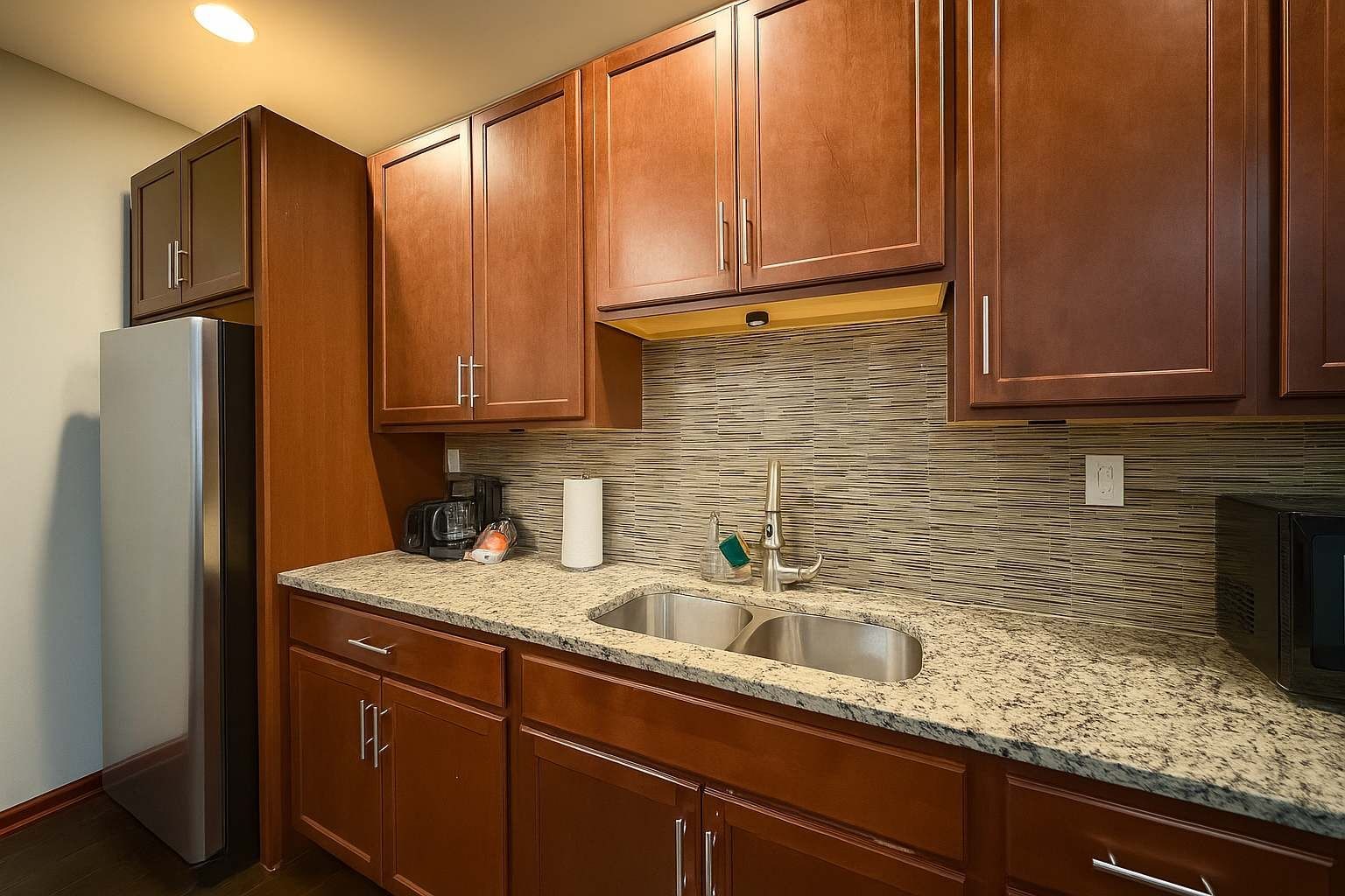 A kitchen with wooden cabinets, a stainless steel refrigerator, granite countertops, and a tiled backsplash.