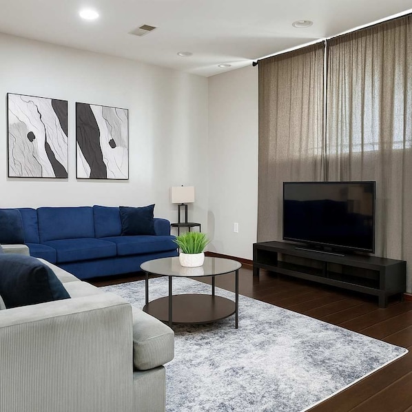 Un salon moderne avec un canapé bleu, un fauteuil blanc, une table basse ronde agrémentée d’une plante, un téléviseur à écran plat et des œuvres d’art abstraites accrochées au mur.