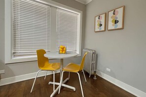 A room with two yellow chairs around a small white table, a radiator, and two framed artworks on the wall. - 1BR Stylishly Apt in Lakeview (Chicago)