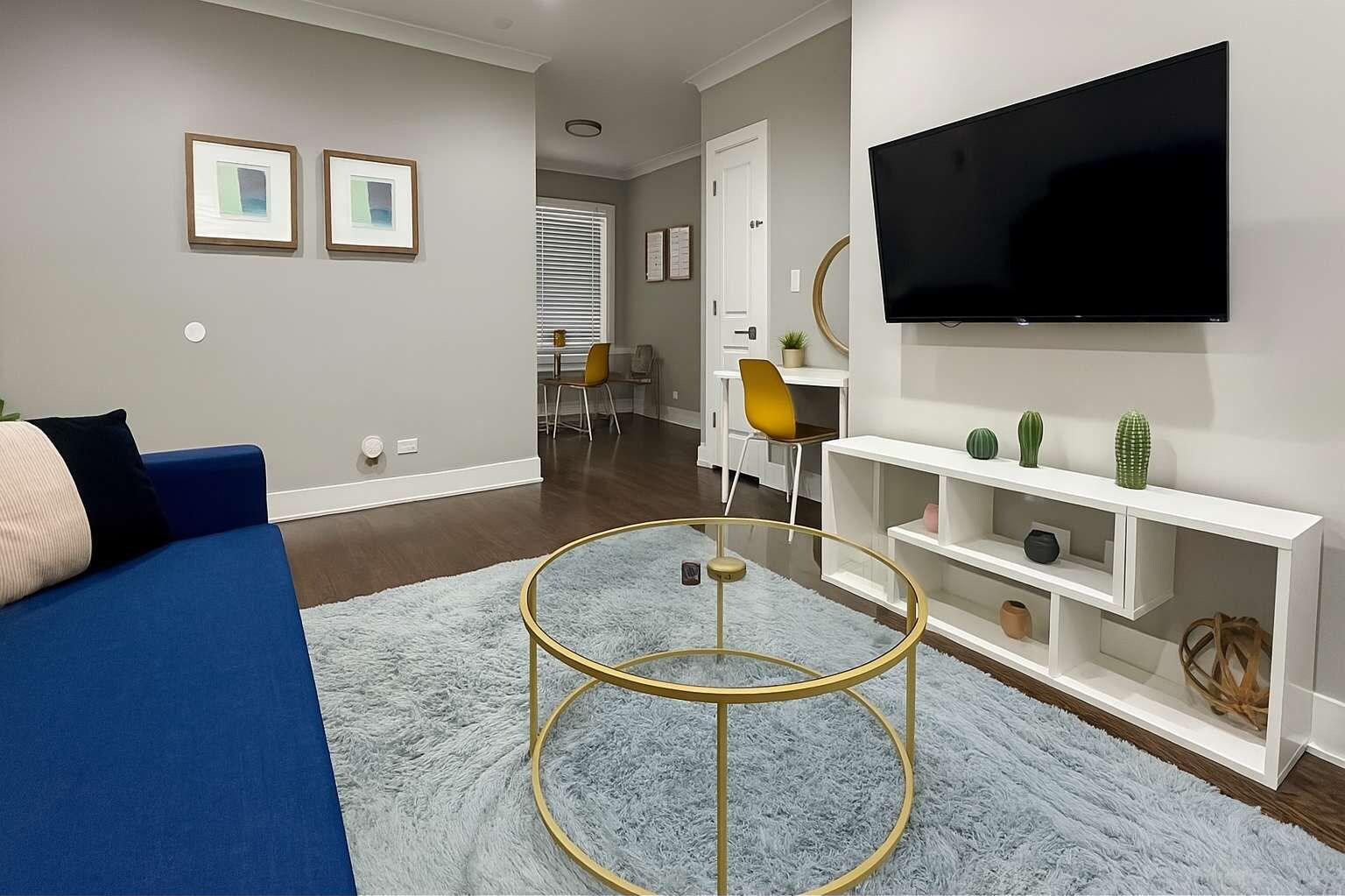 A modern living room with a blue sofa, a round glass coffee table, a flat-screen TV mounted on the wall, and a white entertainment unit with decorative items.