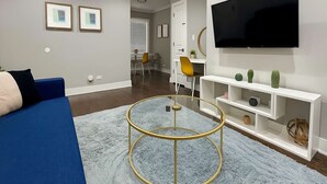 Una sala moderna con un sofá azul, una mesa de centro redonda de vidrio, un televisor de pantalla plana montado en la pared y un mueble de entretenimiento blanco con objetos decorativos.