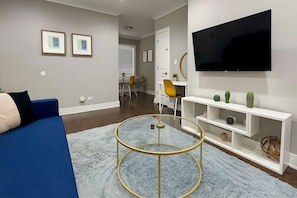 A modern living room with a blue sofa, a round glass coffee table, a flat-screen TV mounted on the wall, and a white entertainment unit with decorative items. - 1BR Stylishly Apt in Lakeview (Chicago)