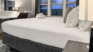 A neatly made bed with white linens, a dark grey headboard, and a stack of white towels on the foot of the bed. There is a window with curtains, a desk with a chair, and a lamp on the side table.