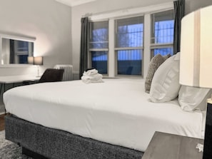 Una cama bien tendida con sábanas blancas, un cabecero gris oscuro y una pila de toallas blancas al pie de la cama. Hay una ventana con cortinas, un escritorio con una silla y una lámpara en la mesita de noche.