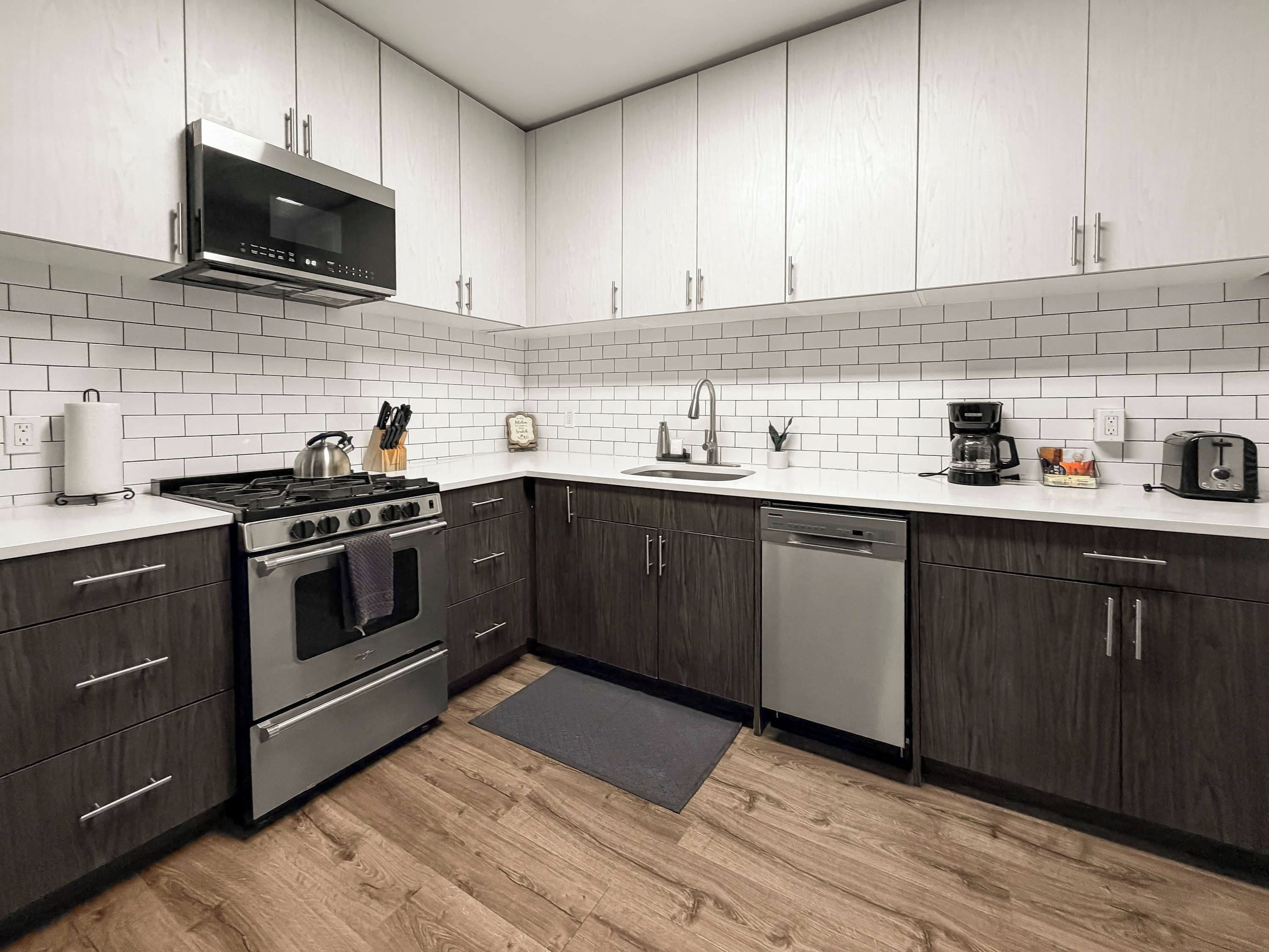 Une cuisine moderne avec des meubles de couleur bois foncé, des carreaux de céramique blancs disposés en motif métro, des appareils électroménagers en acier inoxydable et un plancher en bois.