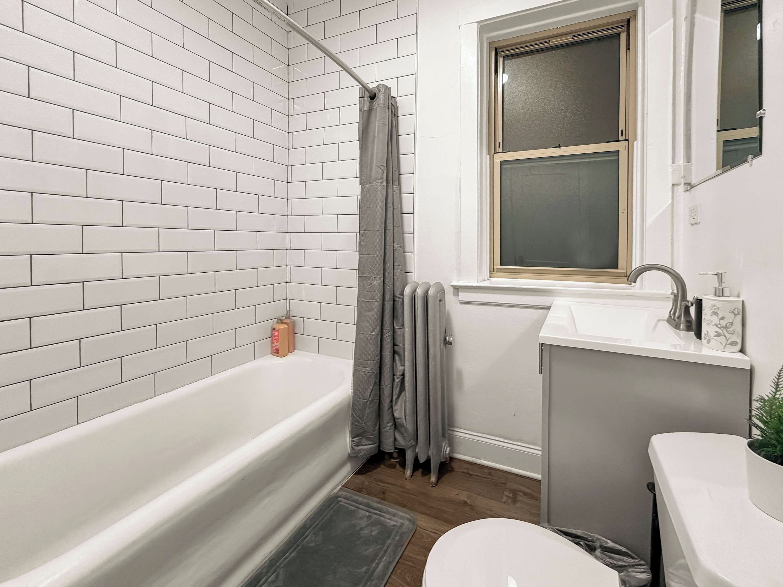 A bathroom with a white bathtub, a window above the sink, and a radiator.