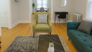 A living room with a green armchair, a teal sofa, a coffee table with a vase and remote control, and a ceiling fan. - Cozy 1BR in Walkable Area (Chicago)