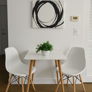 Quarto moderno com mesa branca, duas cadeiras brancas, planta em vaso, pintura abstrata em preto e branco, termostato e uma porta.