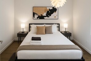 Quarto de hotel bem organizado, com uma cama grande, duas mesas de cabeceira e uma pintura abstrata moderna acima da cama.