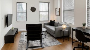 Una sala moderna con un sofá gris, un sillón negro, una mesita redonda con una lámpara, un televisor sobre un mueble bajo y una planta en una mesita auxiliar.