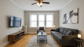 A modern living room with a flat-screen TV, a grey sofa, a coffee table, and a ceiling fan. - Charming 1BR in Old Irving (Chicago)