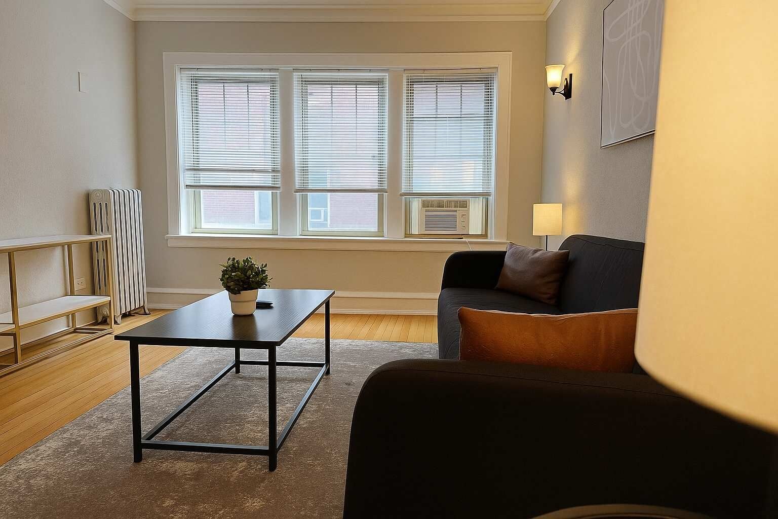 A living room with a dark sofa, a small table with a plant, a radiator, and a window with blinds.