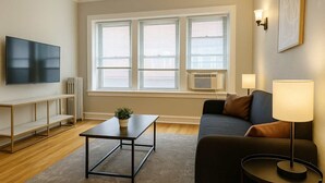 A modern living room with a flat-screen TV, a wooden coffee table, a sofa, a side table with a lamp, and a potted plant.