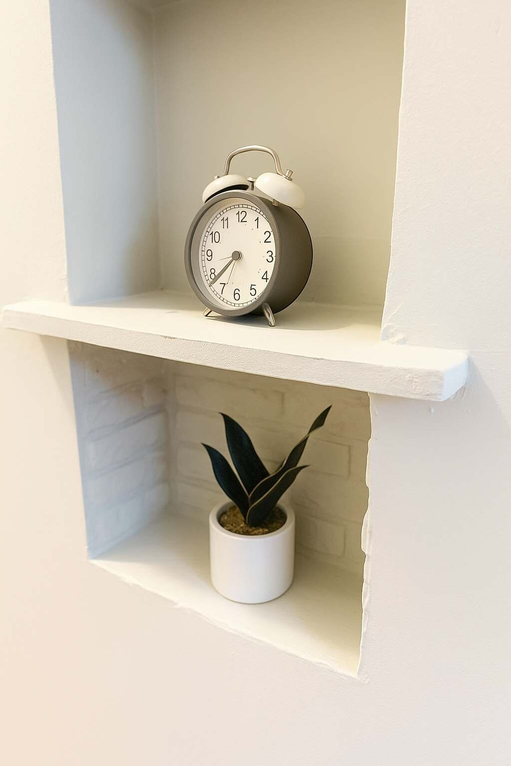 A white shelf with a wall-mounted alarm clock and a potted plant.