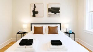 A hotel room with a large bed, two nightstands, two wall-mounted abstract paintings, and a window with blinds.
