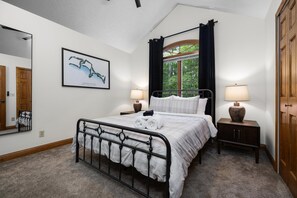 Een slaapkamer met een metalen bedframe, twee nachtkastjes met lampen, een groot raam met gordijnen, een ingelijste foto aan de muur en een deur.