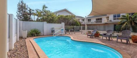 A swimming pool with a curved edge, surrounded by a brick border, and a shaded area with chairs.