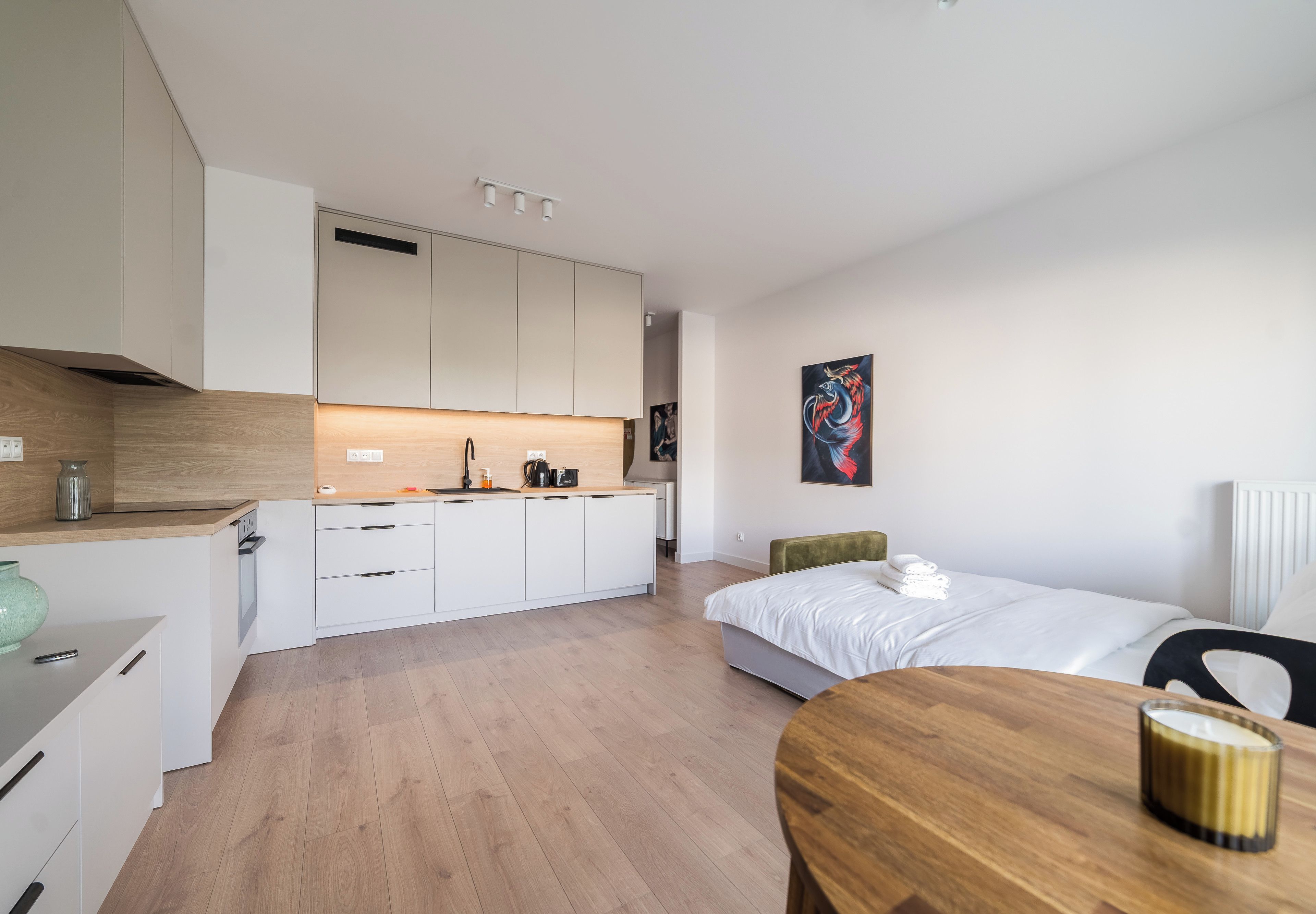 Une chambre d’hôtel moderne dotée d’une kitchenette, d’un lit avec une literie blanche, d’une table à manger en bois et d’un tableau accroché au mur.