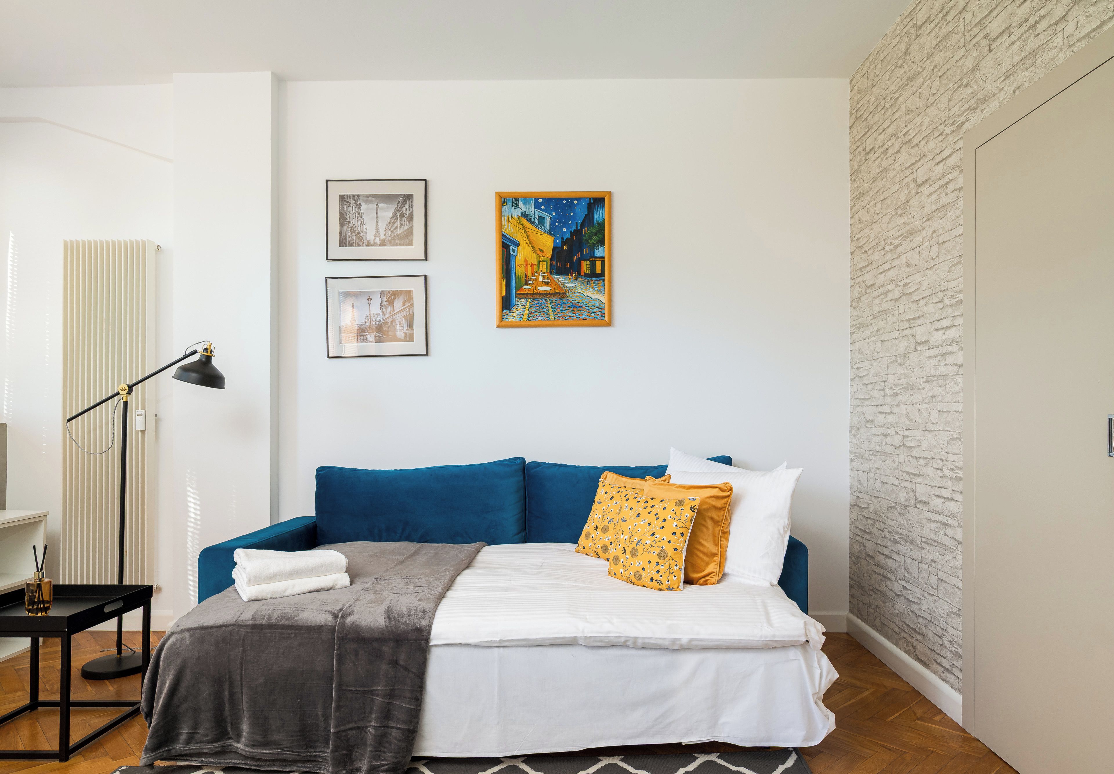 A modern bedroom with a blue sofa, a bed with white and yellow pillows, and two framed artworks on the wall.