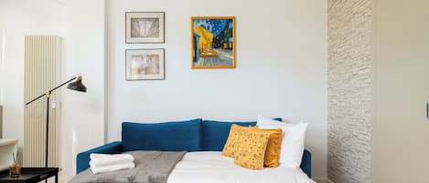 A modern bedroom with a blue sofa, a bed with white and yellow pillows, and two framed artworks on the wall.