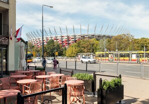 Terrasse mit Tischen und Stühlen, Straßenansicht mit Autos und einer Straßenbahn, und ein großes Stadion im Hintergrund.