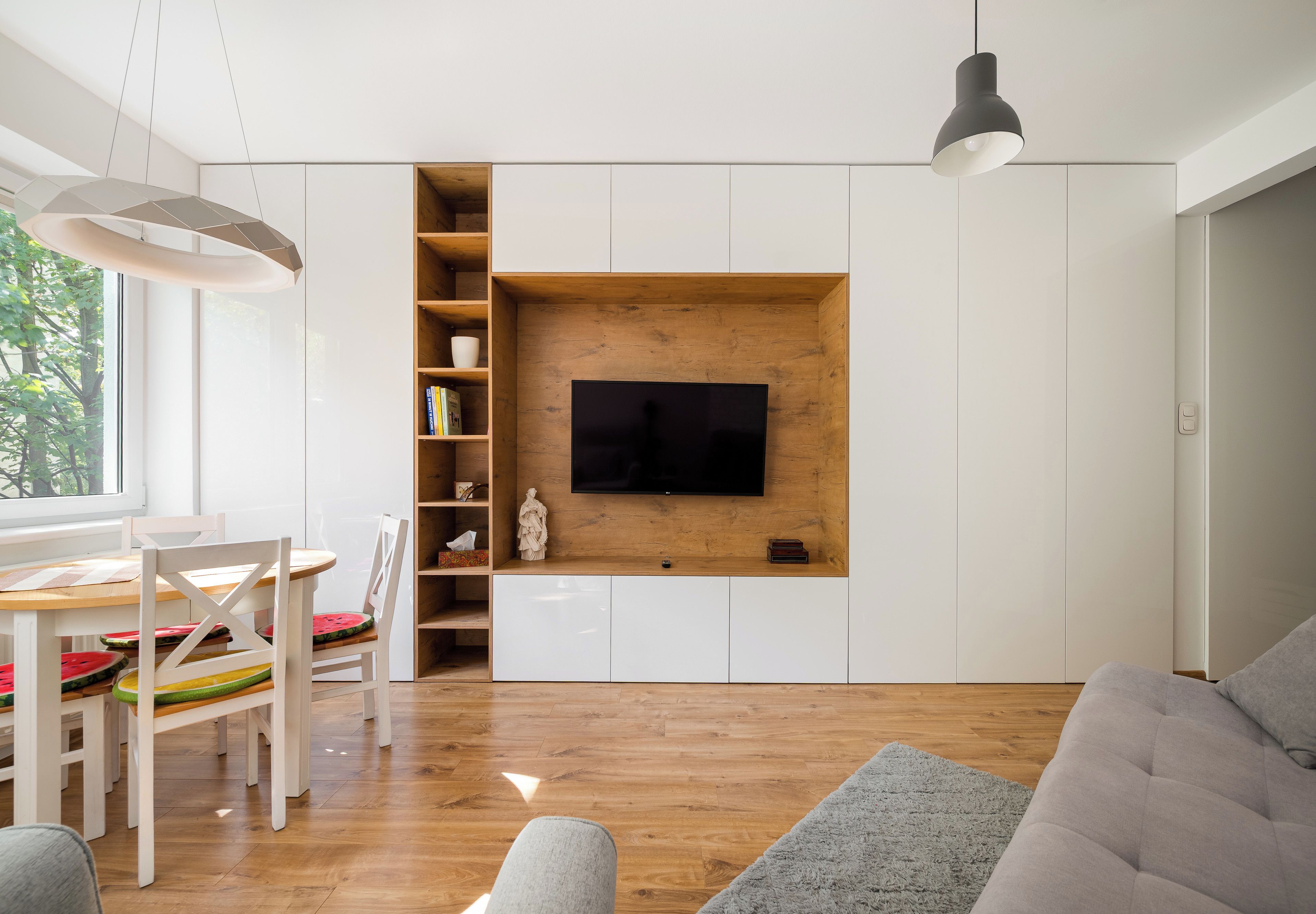 A modern living room with a flat-screen TV mounted on a wooden wall, a dining area with a white table and chairs, and a grey sofa.