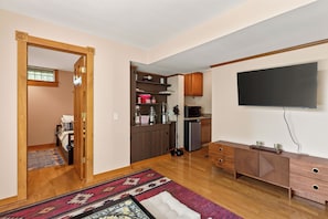 A compact living space with a kitchenette, a flat-screen TV mounted on the wall, and a wooden cabinet with drawers. - Hot Tub | Sauna | Sleeps 6 (Canandaigua)