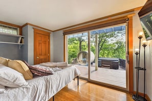 A bedroom with a bed, a wooden door, and a large sliding glass door leading to an outdoor deck with a hot tub and a circular seating area. - Hot Tub | Sauna | Sleeps 6 (Canandaigua)